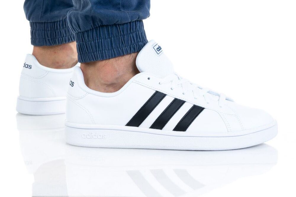 A pair of Adidas men's white sneakers with black stripes, shown on feet with blue joggers, ideal for casual or sporty wear.