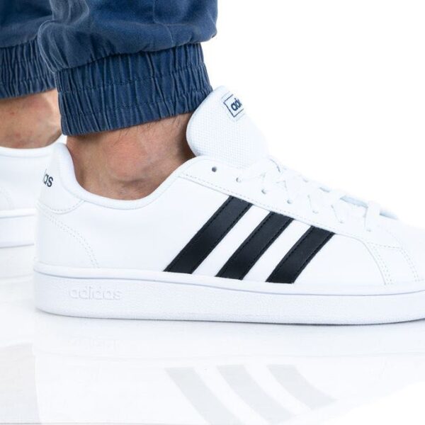 A pair of Adidas men's white sneakers with black stripes, shown on feet with blue joggers, ideal for casual or sporty wear.