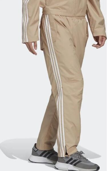 A man wearing Adidas beige track pants with white side stripes, paired with matching beige outerwear and grey sneakers, showcasing a stylish and sporty look.