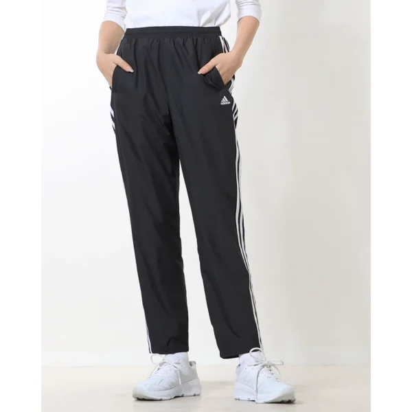Adidas Women's 3-Stripes Woven Pants, Black. Full view, showing black pants with white 3-Stripes down the side, on a person standing with hands in pockets.